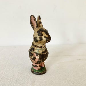 May include: A small, upright rabbit figurine made of cast metal with a weathered, distressed finish. The surface features patches of dark brown, cream, and faded pink paint, revealing the underlying metal texture across the ears, body, and base.