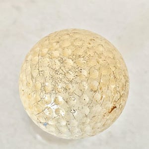May include: A golf ball with a textured surface and a pattern of small dimples. The ball has a light beige color with darker cracks and markings across its surface. The golf ball is a sphere.