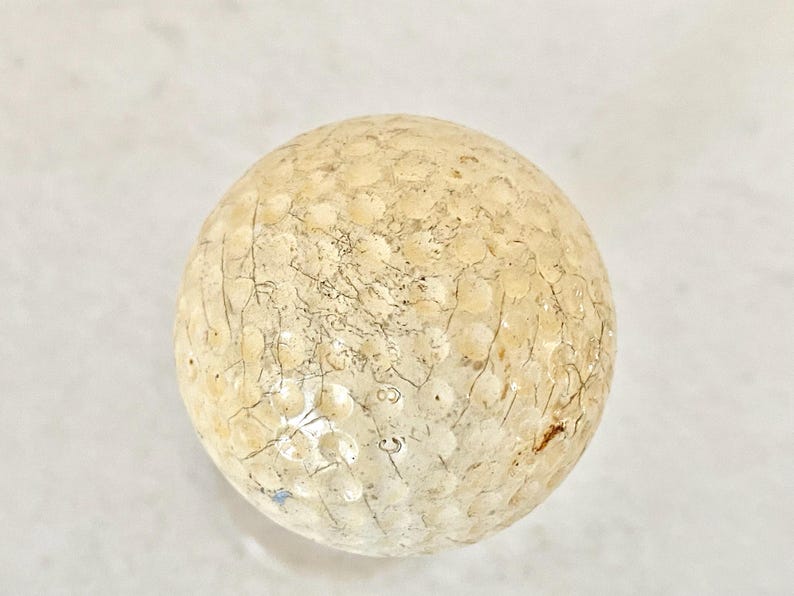 May include: A golf ball with a textured surface and a pattern of small dimples. The ball has a light beige color with darker cracks and markings across its surface. The golf ball is a sphere.