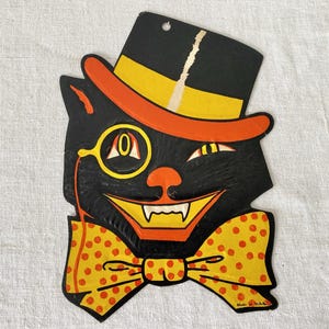 May include: A die-cut cardboard decoration featuring a black cat wearing a black top hat with a yellow band and orange brim. The cat has a yellow monocle, a wide grin with visible white fangs, and a large yellow bow tie with orange polka dots.