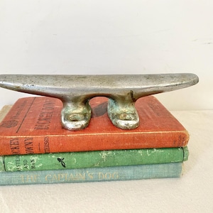 May include: A weathered, silver-toned boat cleat, resting on a stack of vintage books with red, green, and blue covers. The cleat's design features a curved top and two base supports, suggesting nautical decor.