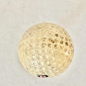 May include: A used golf ball with a dimpled surface. The ball is off-white with brown and black markings and some visible wear. The ball has a red and black logo.