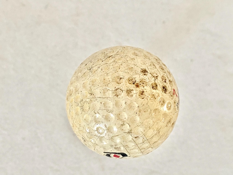 May include: A used golf ball with a dimpled surface. The ball is off-white with brown and black markings and some visible wear. The ball has a red and black logo.