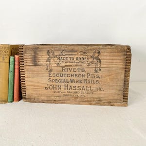 May include: Vintage wooden crate with the text "RIVETS, ESCUTCHEON PINS, SPECIAL WIRE NAILS. JOHN HASSALL, INC." and illustrations. The crate is next to a stack of old books with colorful spines. The crate is made of weathered wood.