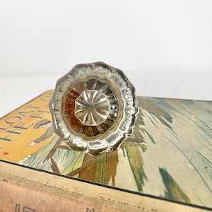 May include: A clear glass door knob with a decorative, ridged edge. The knob is resting on a vintage book with the title "Afloat the Flotilla". The book has a yellow cover with illustrations.