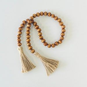 May include: A brown wooden bead garland with two tassels. The garland is made of round wooden beads and has a natural brown color. The tassels are made of a natural fiber and are a light brown color.