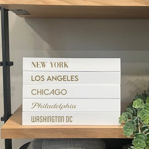 May include: A stack of five white decorative books with gold lettering. The spines are printed with the city names NEW YORK, LOS ANGELES, CHICAGO, PHILADELPHIA, and WASHINGTON DC in various fonts, stacked horizontally on a wooden shelf.