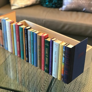 Könnte beinhalten: Ein Bücherregal aus Holz mit einer Vielzahl von bunten Büchern. Die Bücher sind vertikal angeordnet und haben alle unterschiedliche Größen. Das Bücherregal steht auf einem Glastisch.