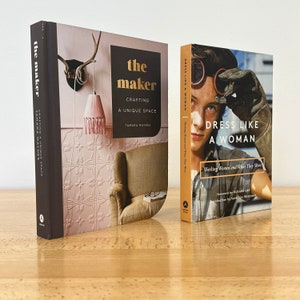 May include: Two hardcover books on a wooden table. The book on the left is titled "The Maker: Crafting a Unique Space" by Tamara Maynes. The book on the right is titled "Dress Like a Woman: Working Women and What They Wore" with a foreword by Roxane Gay and an introduction by Vanessa Friedman.