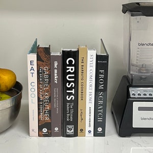 May include: A collection of nine cookbooks with various titles, including "Eat Cool", "Gabriel Kreuther", "The Maker", "Crusts", "The Barbuto Cookbook", "Style Comfort Home", and "From Scratch". The books are arranged vertically on a white countertop.
