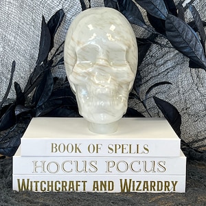 Puede incluir: Un cráneo de cerámica blanca con vetas doradas se encuentra sobre una pila de tres libros blancos. Los libros tienen letras doradas que dicen "Book of Spells", "Hocus Pocus" y "Witchcraft and Wizardry".