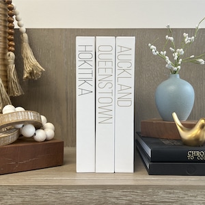 Puede incluir: Tres libros decorativos blancos con letras doradas en los lomos. Los libros se titulan "Auckland", "Queenstown" y "Hokitika".