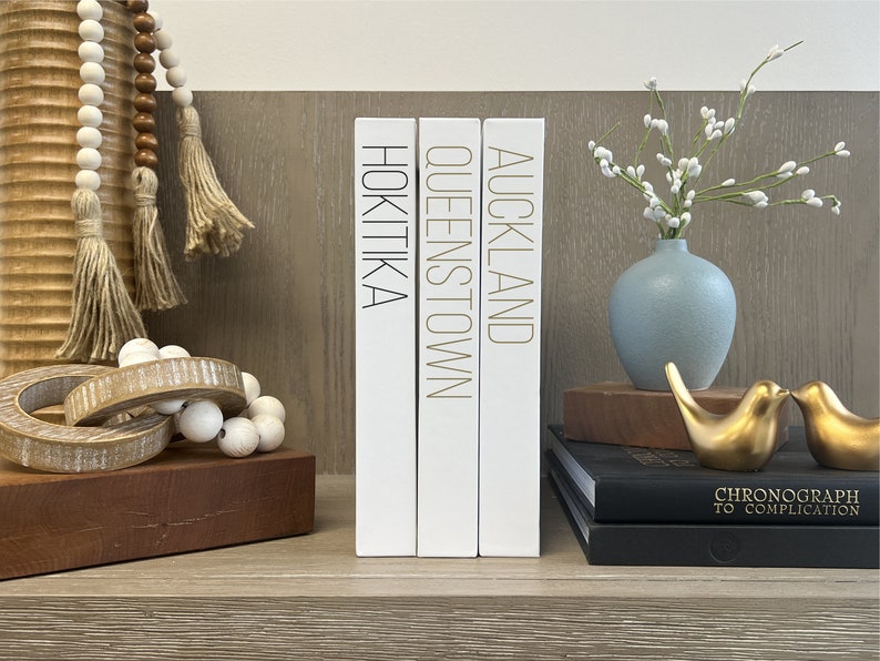 Puede incluir: Tres libros decorativos blancos con letras doradas en los lomos. Los libros se titulan "Auckland", "Queenstown" y "Hokitika".