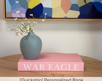 May include: A matte blue ceramic vase containing white berry-like stems sits atop a rectangular pink book with the white text WAR EAGLE printed on the spine. The book and vase are positioned on a round wooden surface with a visible grain pattern.