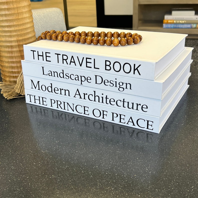 May include: A stack of five white books with black titles. The titles are "The Travel Book", "Landscape Design", "Modern Architecture", "The Prince of Peace", and "The Prince of Peace". A strand of brown wooden beads is on top of the books.