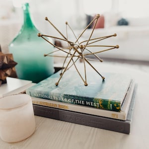 May include: A gold metal decorative stand with a starburst design, a teal glass bottle, a wooden puzzle, and three books stacked on top of each other. The top book is titled 