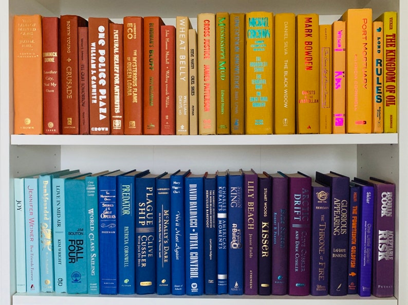 May include: A bookshelf with two shelves filled with a variety of colorful books. The top shelf has books with orange, yellow, and brown spines. The bottom shelf has books with blue, purple, and teal spines. The books are arranged in a rainbow pattern.
