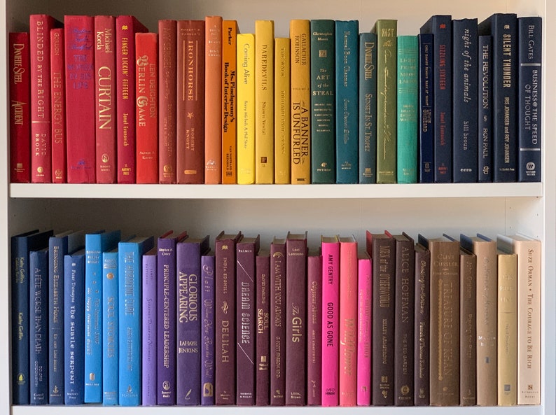 May include: A bookshelf filled with colorful hardcover books. The books are arranged in rows, with the spines facing out. The books are all different sizes and shapes, and they have a variety of titles and authors. The bookshelf is made of wood and has a white finish.