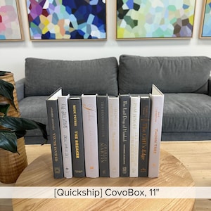 Puede incluir: Una colección de libros de tapa dura con varios títulos y colores, colocados sobre una mesa de madera. Los libros miden aproximadamente 28 cm de altura y se exhiben frente a un sofá gris. El texto "CovoBox, 11" es visible.