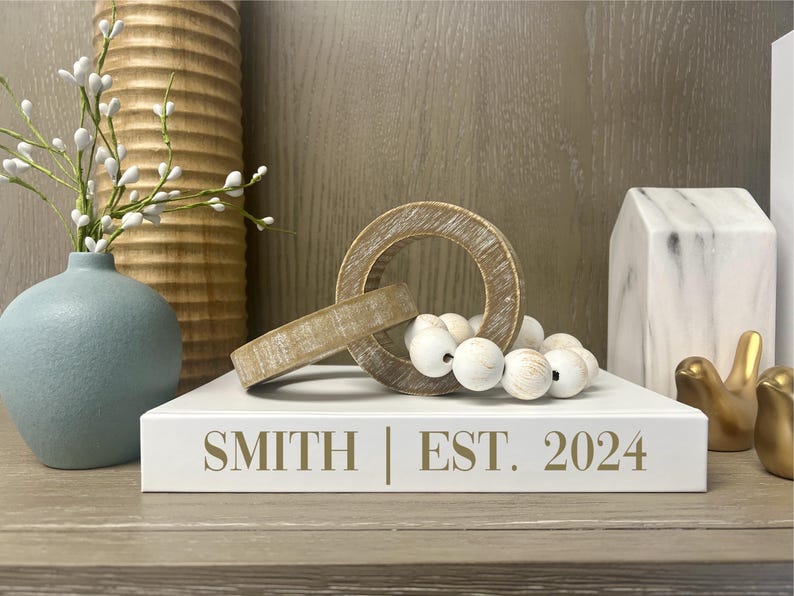 Puede incluir: Libro blanco con el texto "SMITH | EST. 2024" en letras doradas. El libro est&aacute; en un estante de madera con un jarr&oacute;n de madera con flores blancas, un anillo de madera, cuentas blancas, una casa de m&aacute;rmol y dos figuras de p&aacute;jaros dorados.