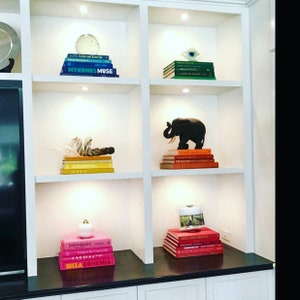 May include: White built-in shelves display colorful stacks of books and decorative objects.  The top shelf features teal and green books, while the second shelf showcases orange and red books.  A black elephant figurine sits atop the orange books.  The third shelf displays yellow and gold books, and the bottom shelf features pink books.  Small decorative items, including a white gourd and a glass orb, add visual interest.  The books' titles are partially visible, including 