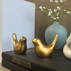 May include: Two gold bird figurines on a black book with the title "Chronograph to Complication". The birds are facing each other and are in a romantic pose.