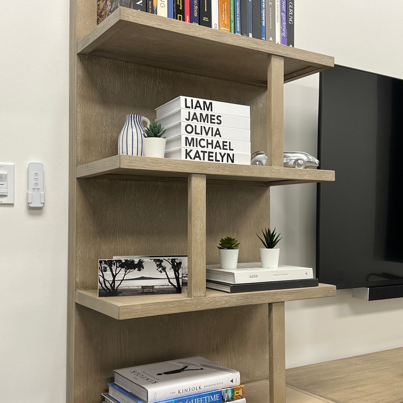 Puede incluir: Una estanter&iacute;a de madera con cinco estantes. El estante superior tiene una fila de libros. El segundo estante tiene una pila de libros blancos con letras negras que dicen "Liam James Olivia Michael Katelyn". El tercer estante tiene una foto en blanco y negro de una escena de playa. El cuarto estante tiene dos peque&ntilde;as plantas en macetas y un libro negro con la palabra "Aero" en la portada. El estante inferior tiene una pila de libros con las palabras "Kinfolk" y "Lifetime" visibles.