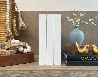 May include: Three white books stacked on a shelf with a wooden surface. The books are all the same size and have a simple, modern design. There are decorative items on the shelf, including a vase with white flowers, a wooden bowl with beads, and a pair of gold birds.