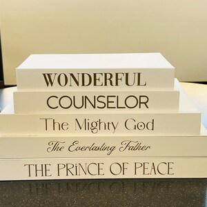 May include: A stack of five white books with brown text. The books are stacked on a table. The text on the books reads: "Wonderful Counselor", "The Mighty God", "The Everlasting Father", and "The Prince of Peace".