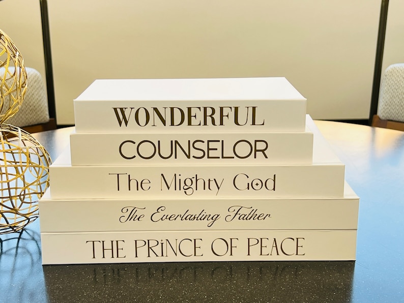 May include: A stack of five white books with brown text. The books are stacked on a table. The text on the books reads: "Wonderful Counselor", "The Mighty God", "The Everlasting Father", and "The Prince of Peace".