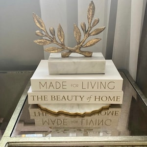 May include: A stack of three books with gold lettering on a glass table. The top book reads "Made for Living", the middle book reads "The Beauty of Home", and the bottom book reads "Made for Living". A gold metal branch sculpture sits on top of the books.