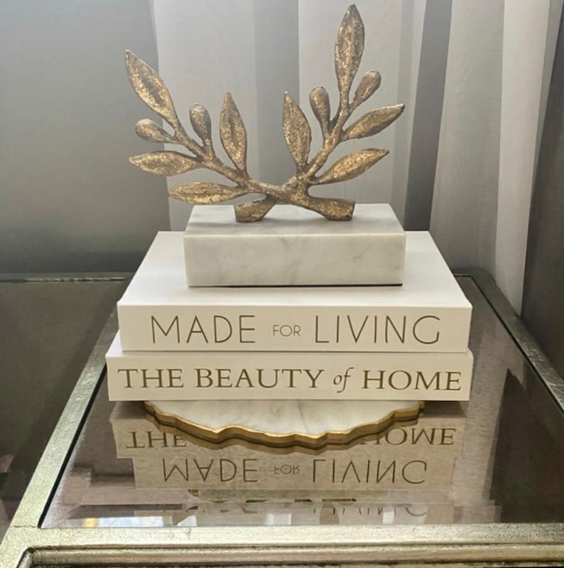 May include: A stack of three books with gold lettering on a glass table. The top book reads "Made for Living", the middle book reads "The Beauty of Home", and the bottom book reads "Made for Living". A gold metal branch sculpture sits on top of the books.