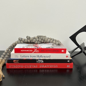 May include: A stack of three books with red covers. The top book is titled "Advanced Love" by Ari Seth Cohen. The middle book is titled "Letters from Hollywood" by Lang & Hall. The bottom book is titled "Strong-Cuevas Drawings" with a red and white bistro chair logo. A gray beaded garland is draped over the top book.