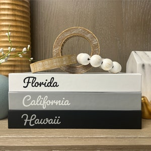 Puede incluir: Tres cajas de madera apiladas con las palabras "Florida", "California" y "Haw&aacute;i" escritas en letra cursiva negra. Las cajas son blancas, grises y negras. Las cajas est&aacute;n sobre una superficie de madera con un anillo de madera, cuentas blancas y figuritas de p&aacute;jaros dorados.