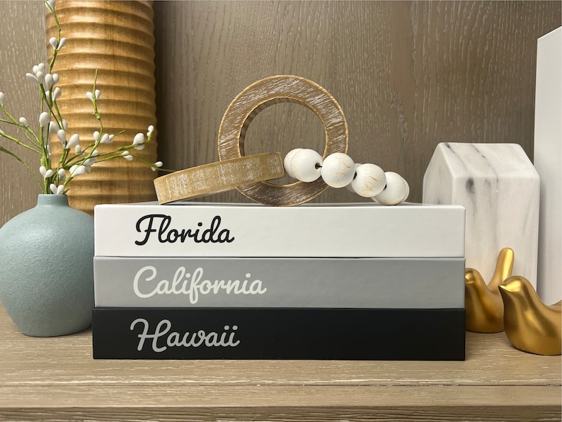 Puede incluir: Tres cajas de madera apiladas con las palabras "Florida", "California" y "Haw&aacute;i" escritas en letra cursiva negra. Las cajas son blancas, grises y negras. Las cajas est&aacute;n sobre una superficie de madera con un anillo de madera, cuentas blancas y figuritas de p&aacute;jaros dorados.