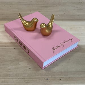 May include: Two metallic gold bird figurines resting on a pink hardback book. The book spine features the text DEBORD in gold, while the front cover displays the names Justin & Camryn in a gold cursive script font against the pink surface.