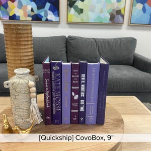 May include: A collection of hardcover books with purple and dark purple spines, arranged on a wooden tray. The books are various sizes and titles, with gold decorative birds and a vase in the foreground. The text "CovoBox, 9"" is visible.