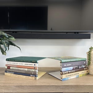 Könnte beinhalten: Auf einer Holzoberfläche sind Bücher mit Titeln wie "Richard Brown's New England" und "Style Comfort Home" gestapelt. Die Bücher sind in zwei Gruppen angeordnet, wobei die Buchdeckel eine brückenartige Struktur bilden. Im Hintergrund ist ein Fernseher zu sehen.
