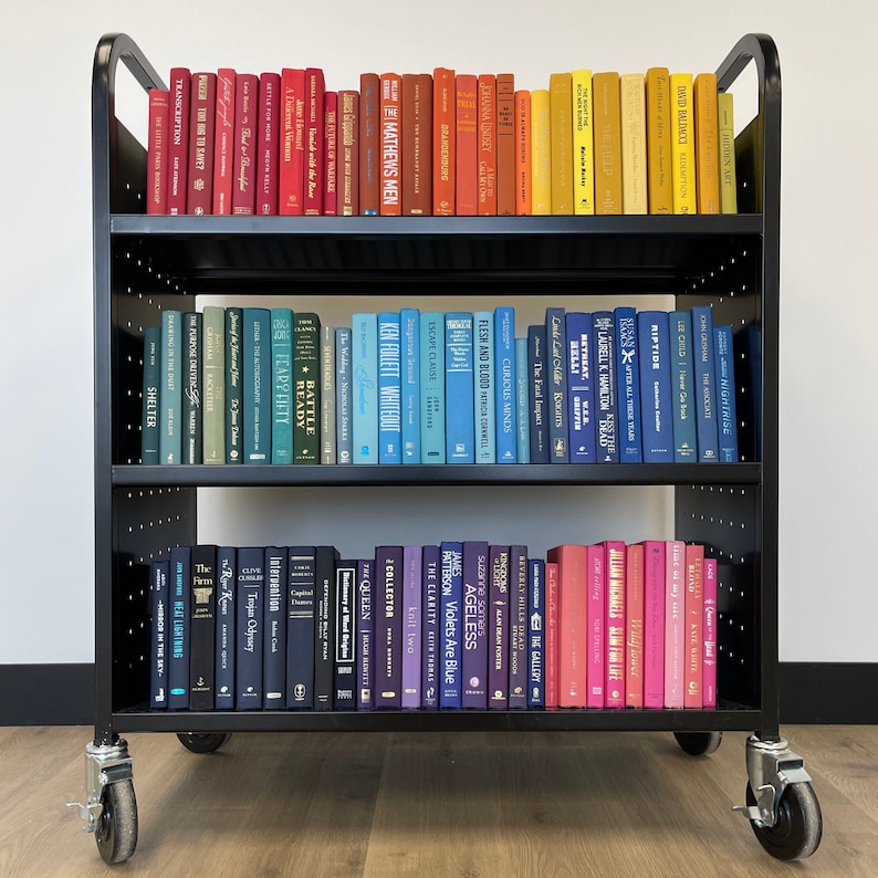 May include: A black metal bookshelf with three shelves filled with various books. The books are organized by color, with red, orange, yellow, blue, and purple books on the shelves. The bookshelf is on wheels and has a white background.