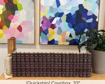 May include: A set of twenty matching dark brown books with gold lettering on the spines, arranged in a single horizontal row. The books feature uniform bindings and are positioned on a wooden surface, with a textured vase and plant nearby.