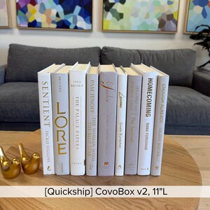 Puede incluir: Una colección de libros de tapa dura con lomos blancos y grises, dispuestos sobre una mesa de madera. Los títulos de los libros son visibles y los lomos tienen letras doradas. Dos figuras de pájaros dorados están en primer plano. El texto "[Quickship] CovoBox v2, 28cm L" está en la parte inferior.