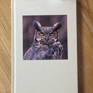May include: A hardcover book with a photograph of a Great Horned Owl on the cover. The owl has yellow eyes and brown and white feathers. The book is white with a clean, minimalist design.