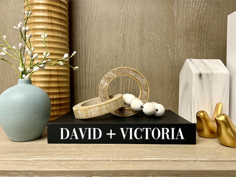 May include: A black book with the text "DAVID + VICTORIA" on the cover. The book is sitting on a wooden shelf with a variety of decorative items, including a blue vase, wooden rings, white beads, and gold bird figurines.
