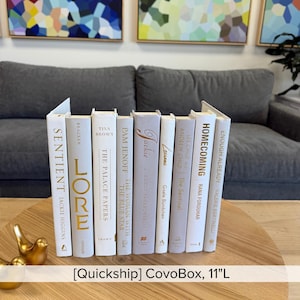 May include: A collection of decorative books in various shades of white and gray, arranged in a row. The books have gold lettering and titles, with the text "[Quickship] CovoBox, 11"L" at the bottom. The books are displayed on a wooden table.