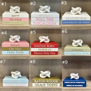 May include: A collection of nine decorative stacks of books, each topped with a white ceramic knot sculpture. The books feature various university-themed text, including SMTTT, Alabama, Hook'em, Rebels, Sooner Born, Columbia, Ole Miss, Baton Rouge, and Move With Purpose.