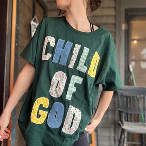 May include: Dark green t-shirt with the words "CHILD OF GOD" appliqued in various patterned fabrics. The letters are in blue denim, floral, gingham, and yellow polka dot fabrics. The shirt is a casual, oversized style.