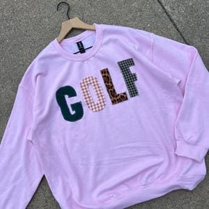 May include: A light pink sweatshirt with the word "GOLF" appliqued across the front. The letters are in various patterns: dark green, gingham, leopard print, and plaid. The sweatshirt is displayed on a wooden hanger.