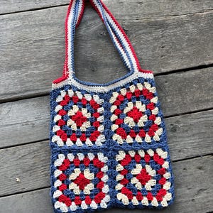 Op de afbeelding: Een handgemaakte gehaakte tote bag met een patchwork ontwerp van rode, witte en blauwe vierkanten. De tas heeft bijpassende rode, witte en blauwe bandjes. De tas is gemaakt van garen en ligt op een houten oppervlak.