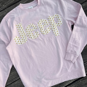 May include: A light pink sweatshirt with the word "Jeep" appliqued across the chest. The letters are made of a white fabric patterned with small yellow ducks. The sweatshirt has a crew neck and long sleeves.
