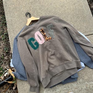May include: A brown sweatshirt with the word "GOLF" appliqued on the front in green, pink gingham, leopard print, and black and white checkered fabric. The sweatshirt is on a wooden hanger, with other sweatshirts and white sneakers in the background.